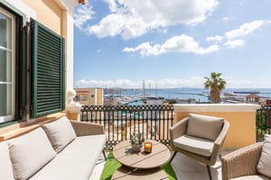 A sunny outdoor terrace featuring wicker seating, a small round table, and green shutters, overlooking a marina and sea.