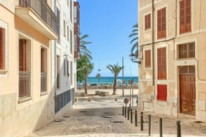 A narrow cobblestone street in a historic Old Town, framed by pale-colored residential buildings with wooden shutters, leading directly to a blue sea view.