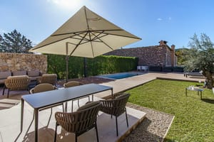 An outdoor dining terrace in Es Capdella featuring a rectangular table with woven chairs, a large beige parasol, and a small pool set against a natural stone wall.