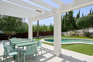 Pérgola blanca sobre mesa de comedor exterior junto a piscina circular con cipreses y setos perfilados