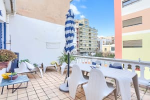 A sunny outdoor terrace with a dining table, chairs, and a blue-striped parasol, featuring city views of neighboring buildings.