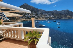 A wide-angle shot of a terrace with white balustrades overlooking the blue waters of Cala Llamp, featuring mountains and anchored boats.