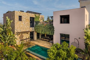 Exterior view of a multi-story villa in Genova featuring a natural stone wall section, smooth pink stucco, a swimming pool, and lush surrounding vegetation.