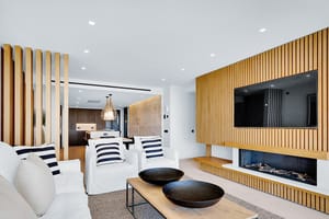 Interior view of a living room featuring a vertical oak slatted wall with an integrated fireplace and TV, white sofas, and coffee table.