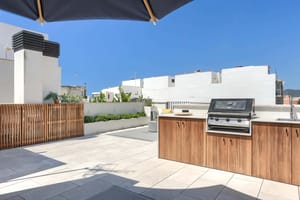 Rooftop terrace featuring a wooden outdoor kitchen cabinet with a built-in stainless steel grill, stone tile flooring, and slatted wood privacy fencing under a clear blue sky.