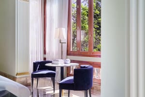 A small circular table with two blue upholstered chairs placed beside tall wood-framed windows with white curtains.