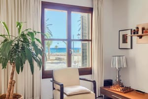 A bright living space with a wooden window frame looking out to the sea and palm trees. An armchair is placed near the window next to a houseplant and a side table with a lamp.