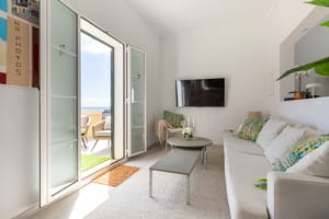 A bright living room with white walls, a beige sofa, and glass doors opening onto a terrace with a glimpse of the sea.