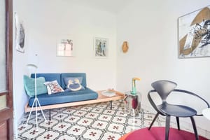 A bright living space featuring a blue sofa, a dark designer chair, and traditional patterned floor tiles.