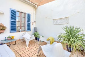 A small outdoor terrace with terracotta tiles, white plastered walls, blue wooden shutters, and modern white lounge chairs.