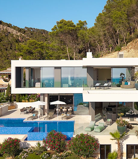 Aerial view of a white, multi-level modern villa built into a hillside, featuring a blue tiled infinity pool and sun loungers.
