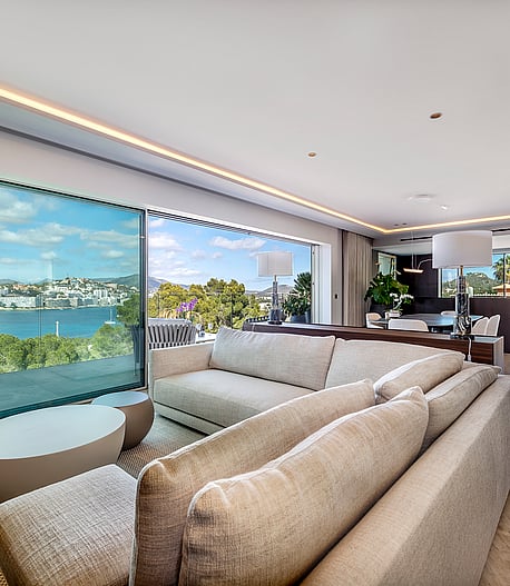 A contemporary open-plan living room featuring a large beige sectional sofa, polished stone floors, and floor-to-ceiling glass sliding doors opening to a lush garden view.
