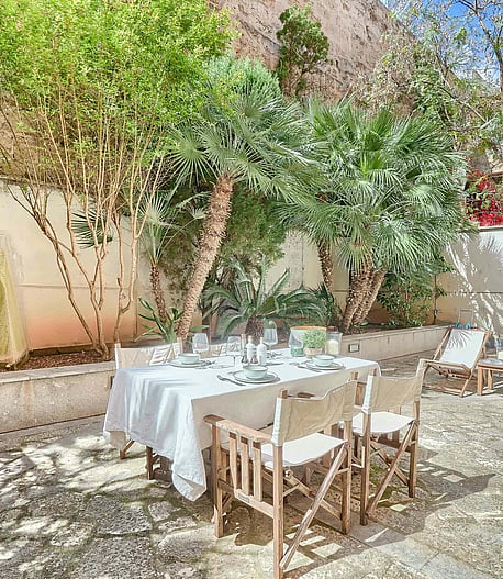 An outdoor dining table with white tablecloth and director chairs set on a gravel terrace shaded by mature trees.