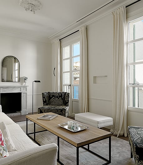 Bright living room with cream furnishings, wooden fireplace mantle, tall windows overlooking the city, and herringbone wooden floors