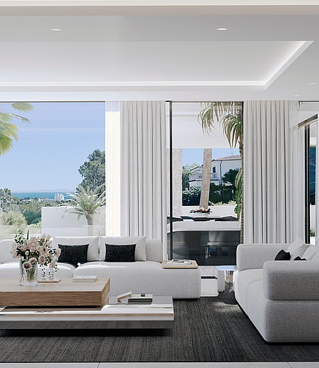 Modern open living room featuring white sofas, a light wood coffee table, dark area rug, and floor-to-ceiling sliding glass doors opening to a covered terrace with coastal views.