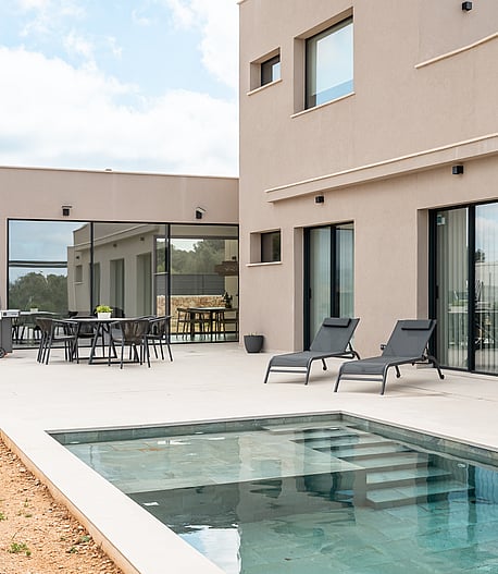Rear facade of a modern beige villa showing a stone terrace, swimming pool with steps, and outdoor dining area.