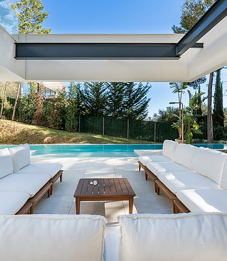 White cushioned outdoor seating under a steel pergola roof beside the swimming pool and pine garden