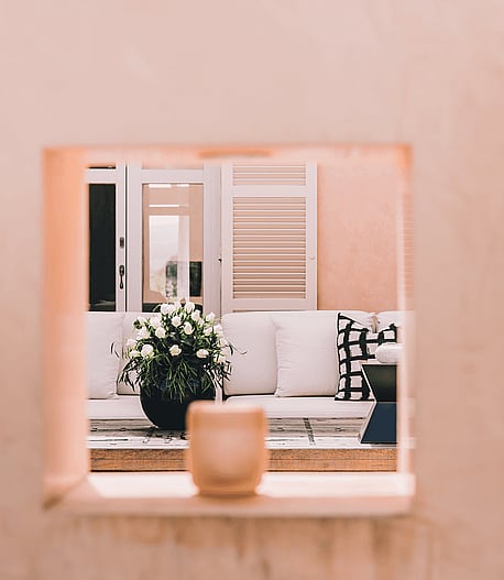 A square architectural opening in a pink stucco wall reveals a white sofa with patterned pillows and a potted plant on a terrace.