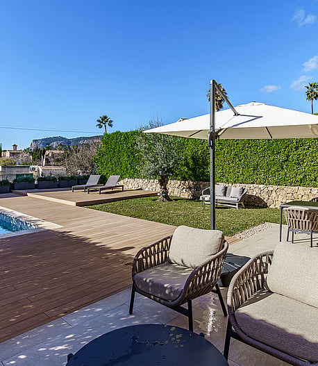 A backyard garden with a small swimming pool, wooden decking, a dining set under an umbrella, and woven outdoor lounge chairs.