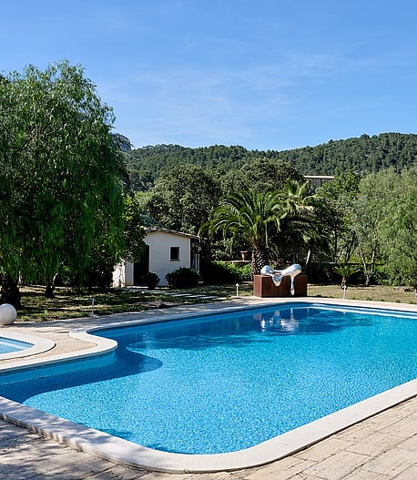 A blue swimming pool with an attached circular spa area, surrounded by a light-colored stone terrace and mature trees against a mountain backdrop.