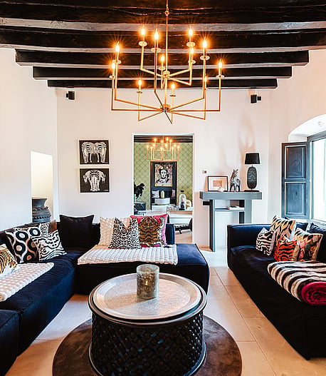 Interior view of a living room with dark low-profile sofas, a central round metal coffee table, and an ornate brass chandelier hanging from dark exposed ceiling beams.