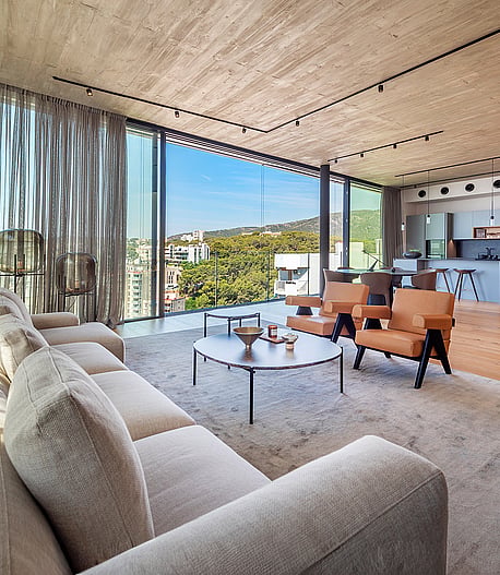 Wide-angle shot of a bright, open-plan living room with exposed concrete ceilings, light wood floors, and floor-to-ceiling glass walls looking out to coastal scenery.