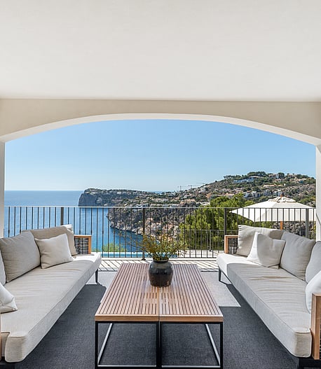 A covered outdoor lounge featuring gray sofas and wooden tables under white arches, overlooking the blue sea and cliffs.