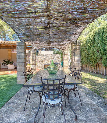 A stone-columned patio with a trellis roof and iron dining set, overlooking a green garden and building exterior.