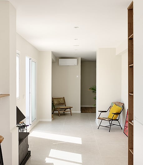 Interior view from a kitchen looking towards a living area with white walls, tiled floors, a stool, and a yellow chair.