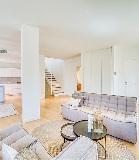 Modern open-plan living room with light oak flooring, grey tufted sofas, and a white architectural staircase.