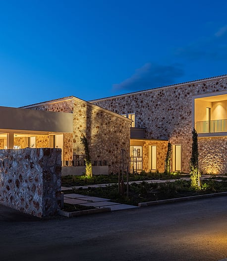 Exterior view of a modern stone-clad finca in Santanyí at twilight, showing illuminated interior spaces and landscaped grounds.