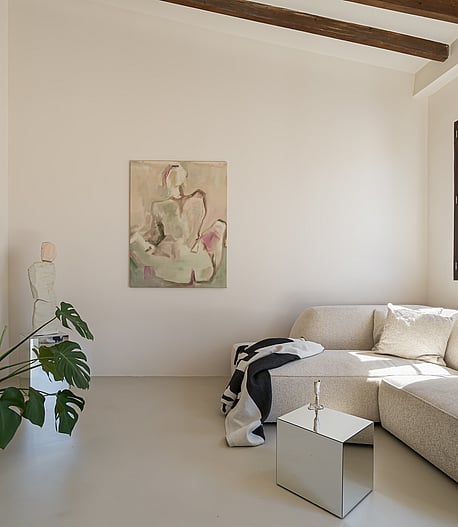 Minimalist living area featuring microcement flooring, light textured sofa, exposed timber beams and a large potted plant.