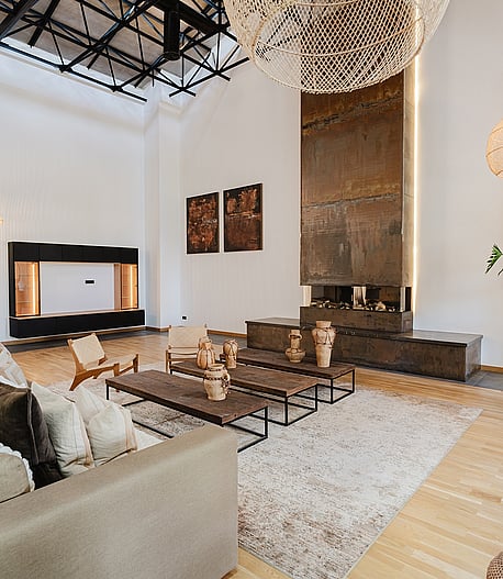 A spacious living area featuring a double-height ceiling with exposed black metal trusses, a tall rusted-metal fireplace surround, and woven pendant lighting.