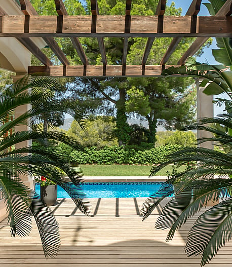 A wooden pergola shades a terrace overlooking a rectangular blue swimming pool with lush garden foliage in the foreground.
