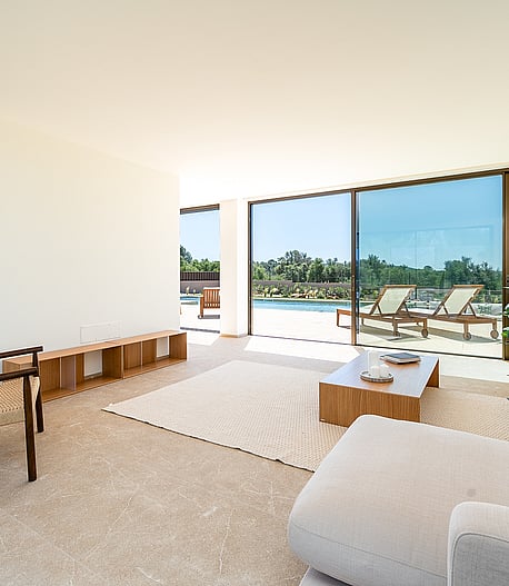 Bright open-plan living room featuring neutral sofas, a woven wood armchair, and floor-to-ceiling glass doors opening to a pool deck.