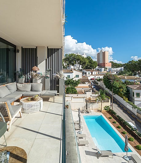 A covered terrace with grey sectional seating overlooks a rectangular swimming pool surrounded by a paved garden in a suburban setting.