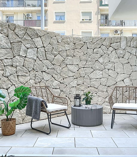 A private outdoor patio featuring a high dry-stone boundary wall, light-toned stone paving, and two rattan chairs with a small table.
