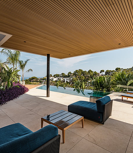 A covered terrace with a wood-slatted ceiling, limestone flooring, and blue outdoor lounge furniture overlooking a swimming pool.