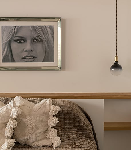 A close-up of a bedroom interior showing a textured brown bedspread, off-white decorative pillows with tassels, and a floating light-oak headboard shelf with a pendant light.