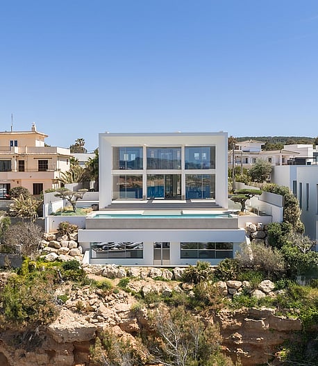 Wide exterior view of a modern white multi-level villa built into a rocky coastline with a swimming pool and glass walls.