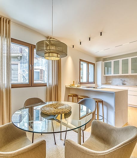 Modern open-plan kitchen with white cabinetry and dining area featuring a round glass table, beige armchairs, and floor-length curtains.