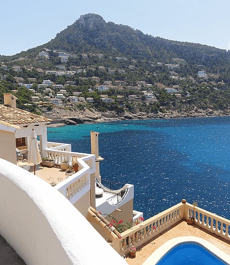 High angle shot of a multi-level stone terrace overlooking a turquoise Mediterranean bay with sun reflecting off the water.