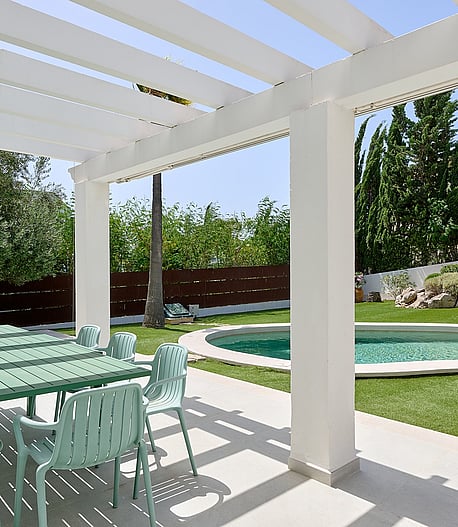 Pérgola blanca sobre mesa de comedor exterior junto a piscina circular con cipreses y setos perfilados