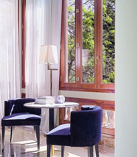 A small circular table with two blue upholstered chairs placed beside tall wood-framed windows with white curtains.