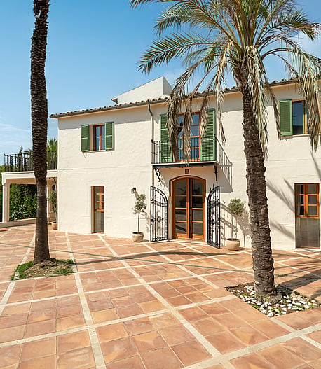 Cream stucco villa with green shutters and an arched wooden door opening onto a geometric terracotta tile courtyard.