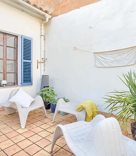 A small outdoor terrace with terracotta tiles, white plastered walls, blue wooden shutters, and modern white lounge chairs.