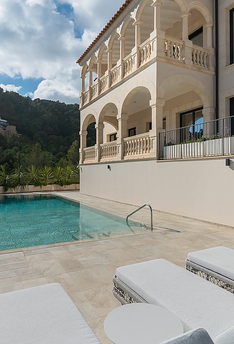 A rectangular swimming pool on a tiled terrace with white sun loungers, set against a multi-story villa with arched balconies.