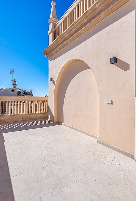 A rooftop terrace featuring light-colored stone flooring, stucco walls with a decorative arch, and stone balustrade railings under a clear blue sky.