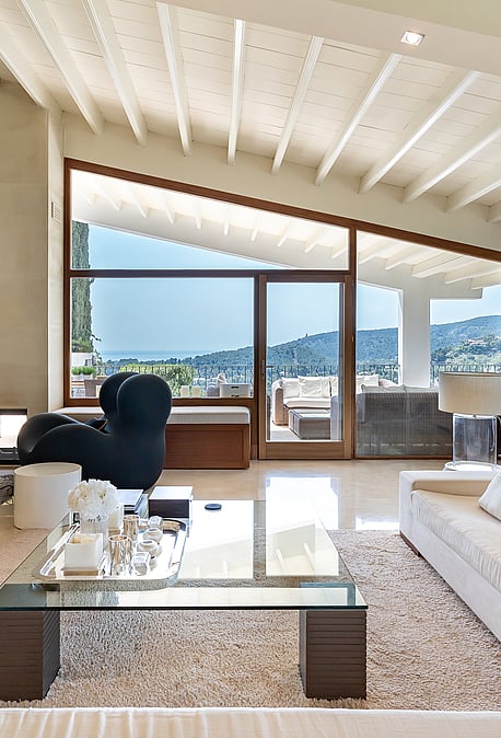 Spacious villa living room with white painted beams, fireplace, marble floors, designer seating and glazed wall with sea views