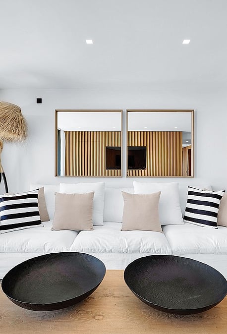 Modern living room featuring a white sofa with striped cushions, two black decorative bowls on a wooden coffee table, and vertical wood slat partitions.